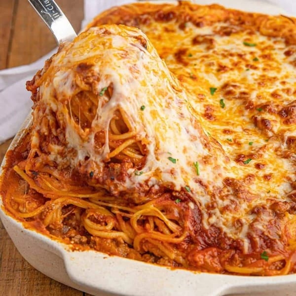 A serving spoon lifts a cheesy portion of baked spaghetti with marinara sauce from a white casserole dish.