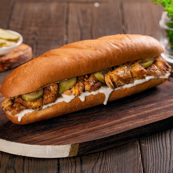 Chicken sub sandwich with pickles and white sauce on a wooden board.